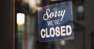 A vintage "Sorry We're Closed" sign on a glass door reflects street view outside.
